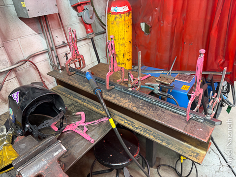 A welding table with a piece of angle iron clamped to a bar with vise-grip C clamps